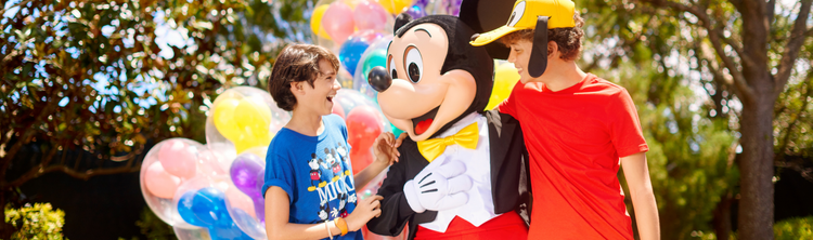 Two boys share a laugh with Mickey Mouse beside a cluster of Mickey balloons.